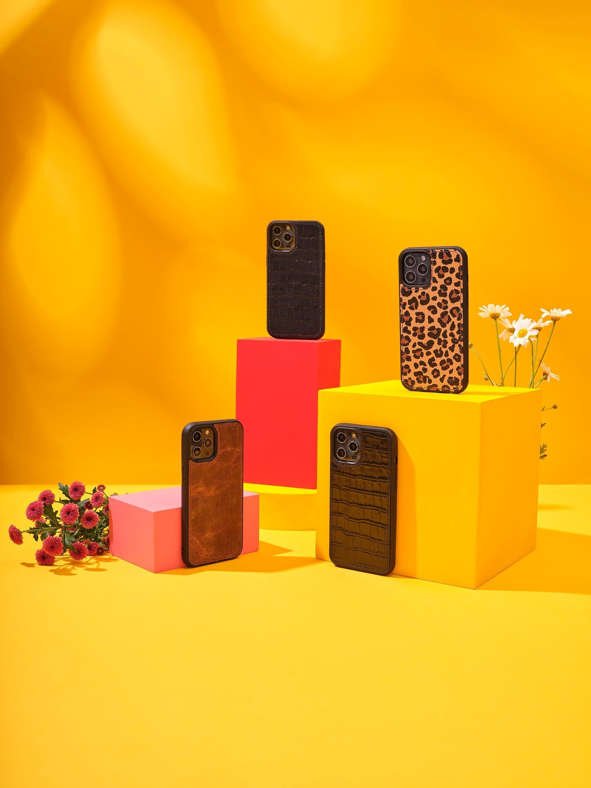 Four leather iPhone cases displayed on colorful blocks against a yellow background, with flowers and studio lighting.