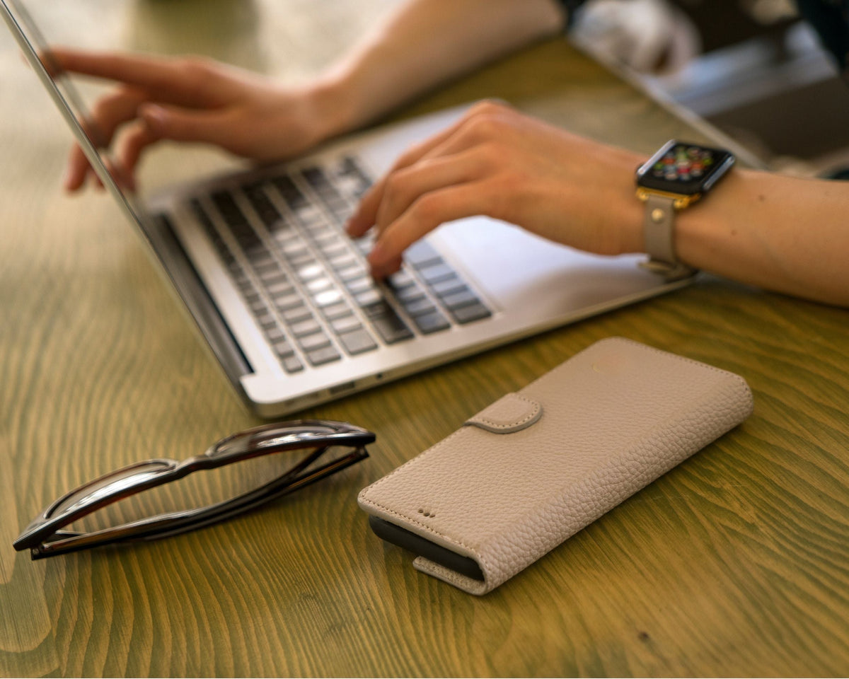 Beige leather wallet phone case beside glasses and a laptop on a wooden desk