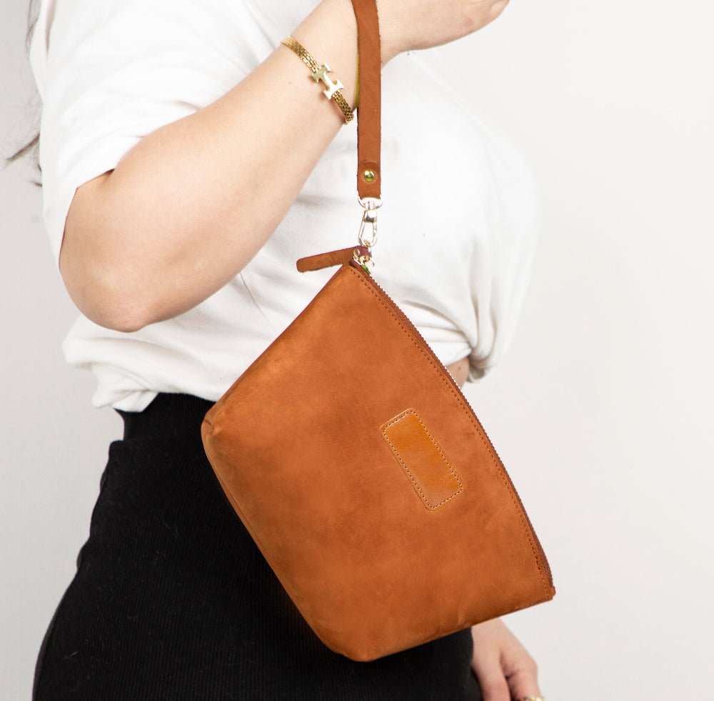 Brown leather crossbody bag held by a person wearing a white shirt and black pants on a light gray background.