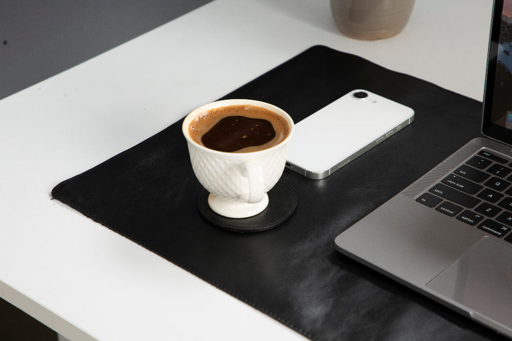 Cup of coffee on a black desk mat with a smartphone and laptop.