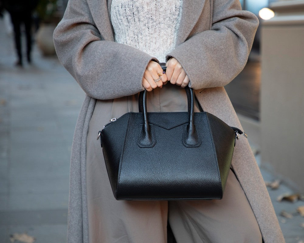 Person holding a black handbag wearing a long coat on a blurred street background