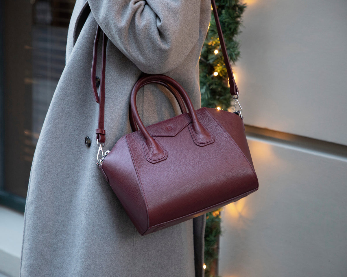 Maroon handbag held by a person wearing a gray coat, with a blurred festive background.
