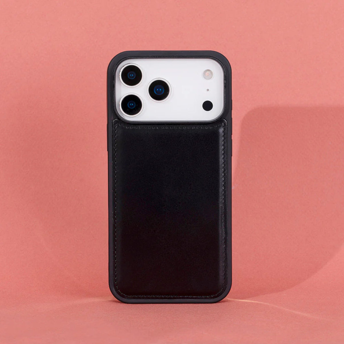 A black leather phone case with a built-in card slot pocket, shown on a white smartphone against a solid pink background.