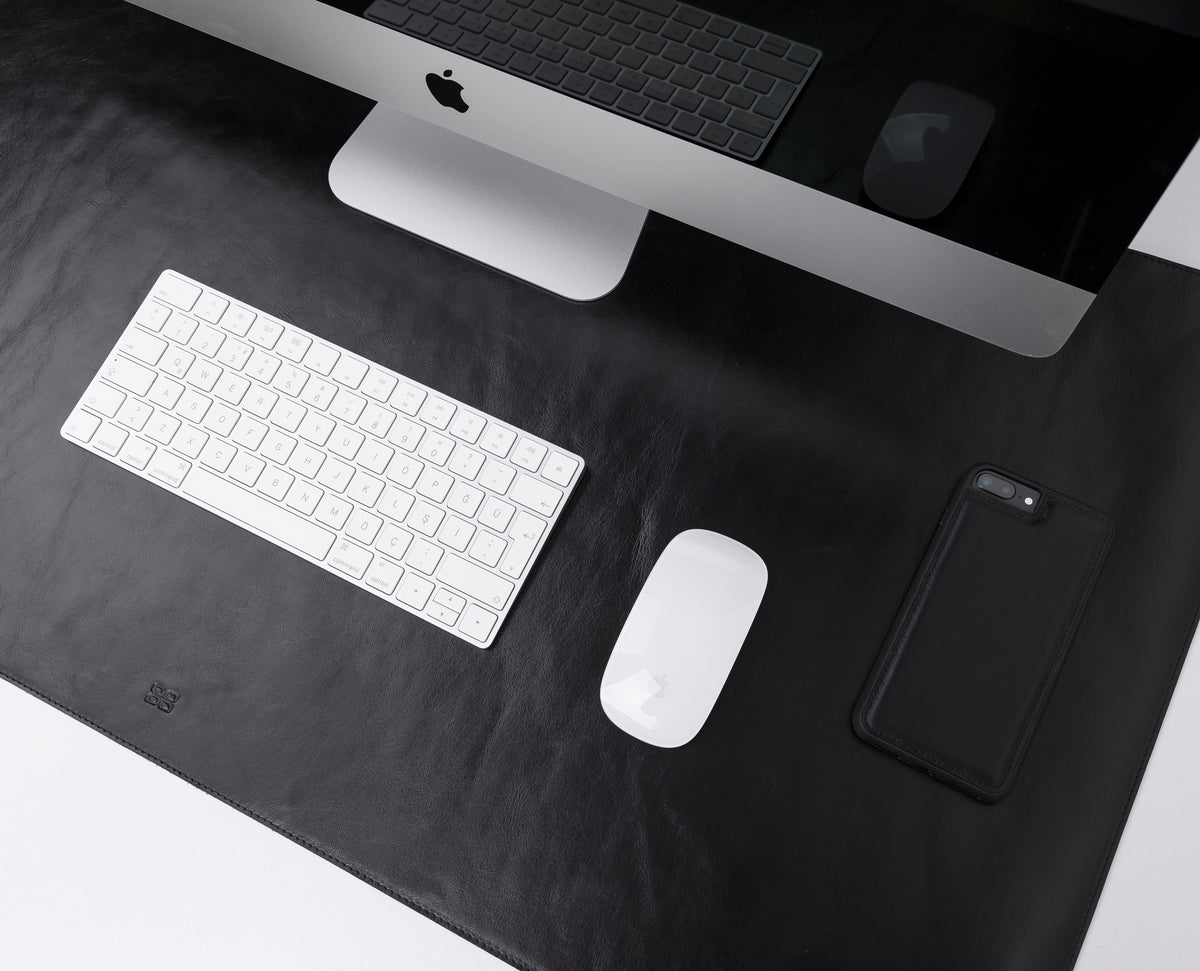 A black genuine leather desk mat on a desk, with a keyboard, mouse, and a smartphone on top of it.