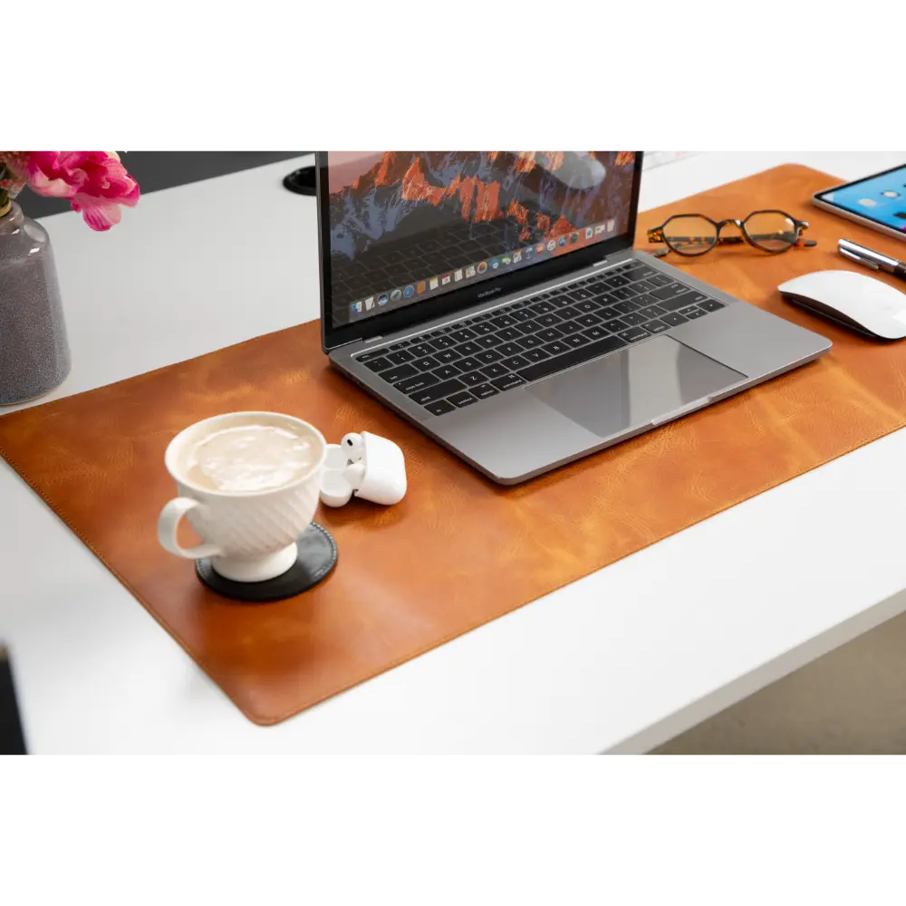 Laptop on a brown leather desk pad with a cup, earbuds, and glasses on a white surface.