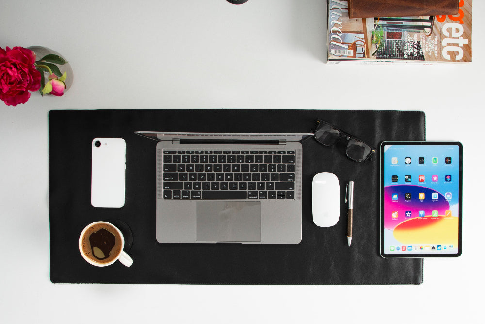Black desk mat with laptop, tablet, phone, and coffee cup on a white surface