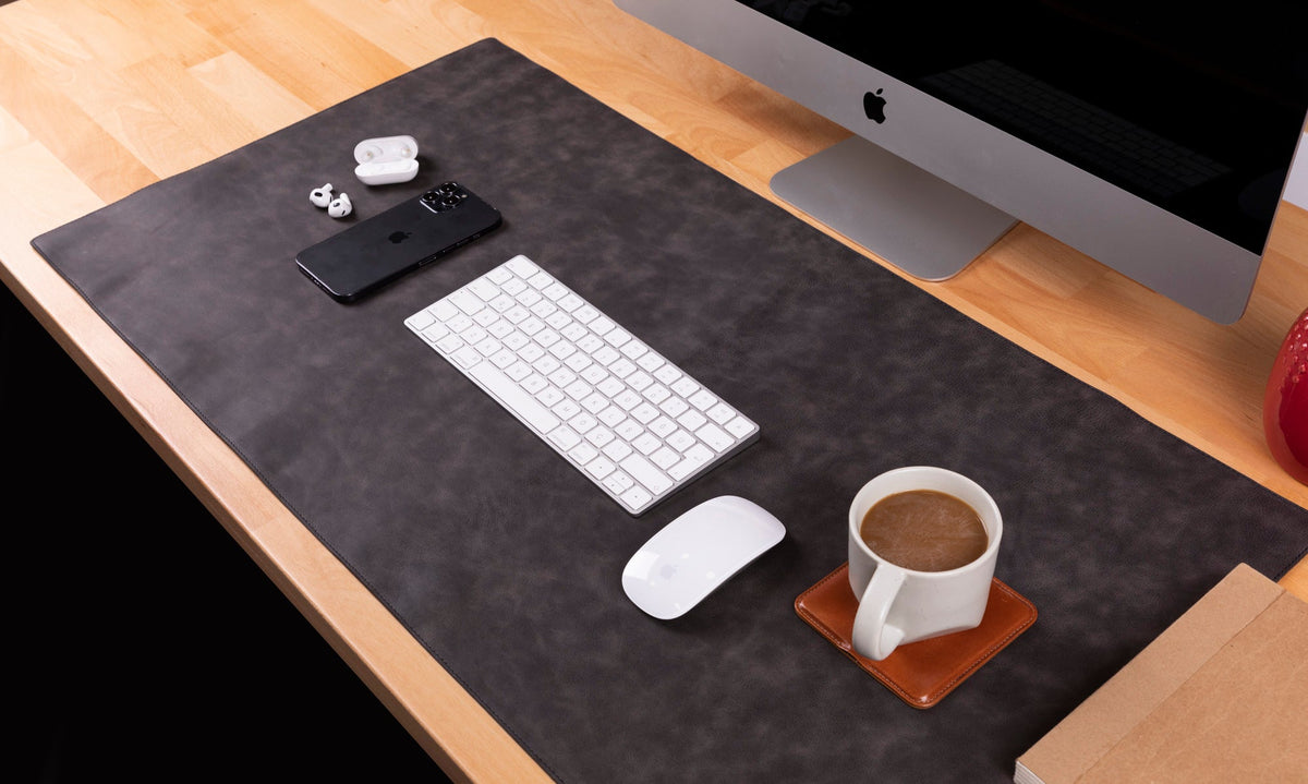 Desk setup with a computer monitor, keyboard, mouse, phone, earbuds, and a cup of coffee on a large desk pad.