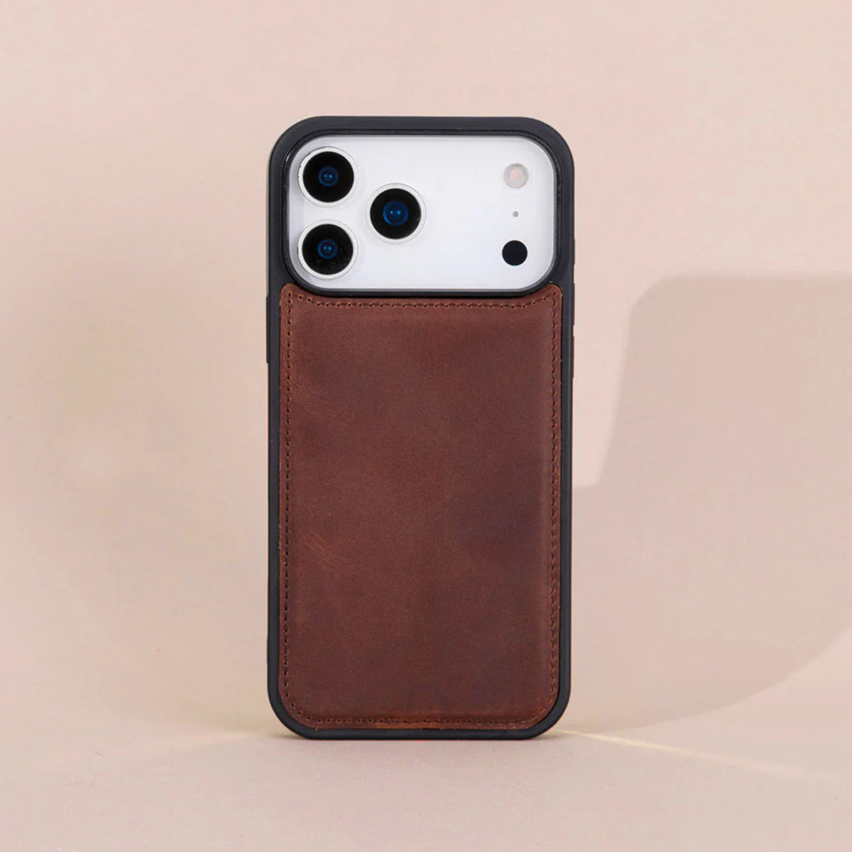 A brown leather phone case with a black bumper frame, shown on a white smartphone against a warm beige background.