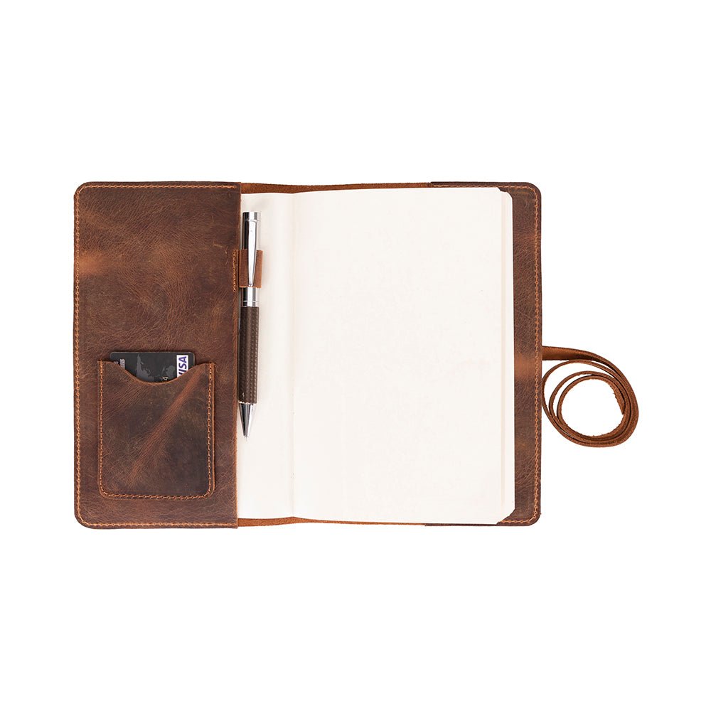 A brown genuine leather journal cover with a pen loop, interior pocket, and wrap-around leather strap closure, displayed open to show the fit of a standard A5-sized notebook.