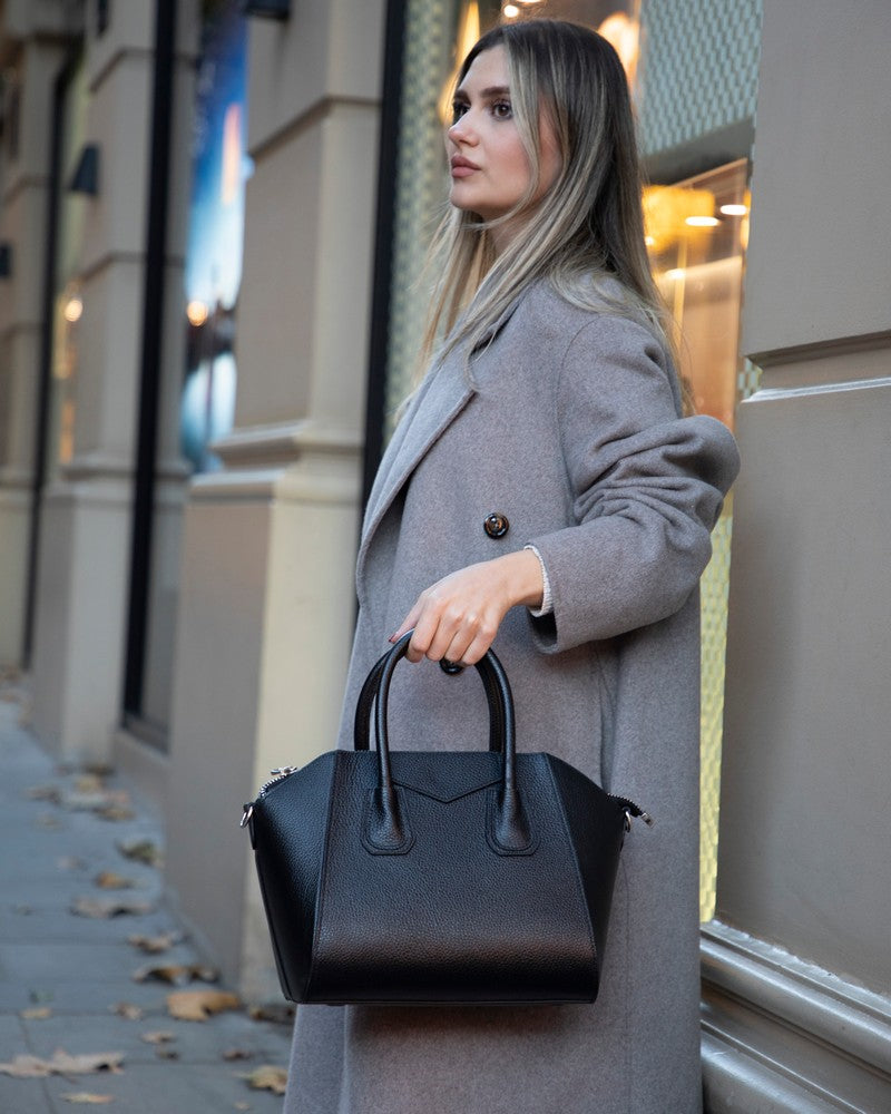 Woman in a gray coat holding a black handbag on a city street.