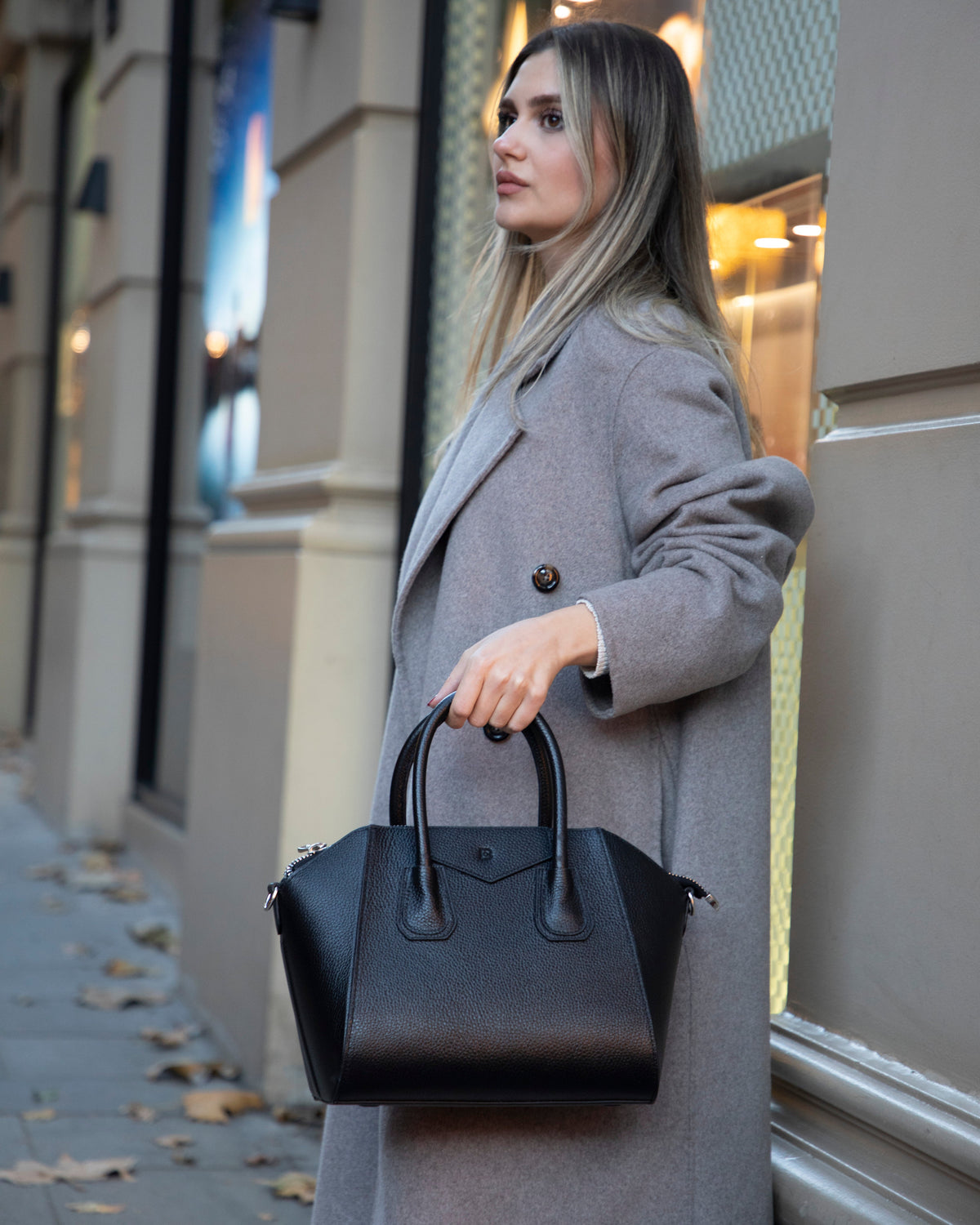 Woman in a gray coat holding a black handbag on a city street.