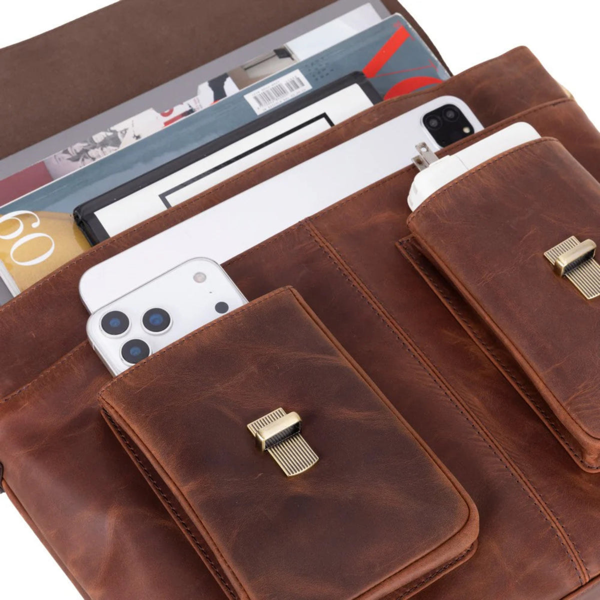 A brown leather organizer pouch or document holder shown open, with multiple compartments holding magazines, cards, and white smartphones. The pouch has two front flap pockets with gold-tone metal clasps and a warm brown leather finish.